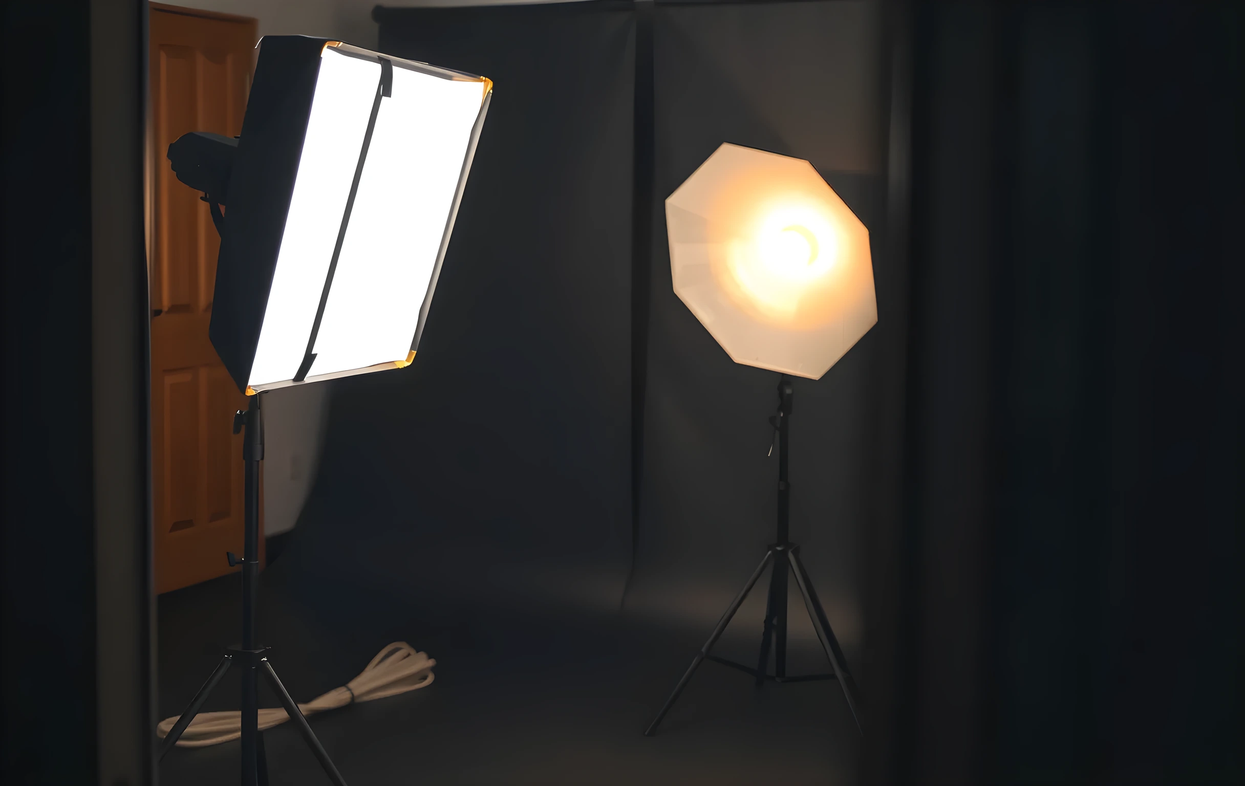 A close detail of a film set lighting rig: a large rectangular softbox hangs from a black C-stand on the left, its diffused panel glowing warm white, behind it a second stand holds a reflector disc angled downward, the background is a deep charcoal cyclorama wall with faint texture, a coiled cable runs along the dark floor in the lower corner, shallow depth of field keeps the softbox edge sharp while the background falls into soft blur, no faces, no screens, no text visible
